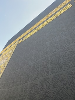 A close-up view of an intricately patterned black and gold surface, featuring Arabic calligraphy with geometric designs. The golden text stands out prominently against the dark background, with a clear sky visible in the upper part of the image.