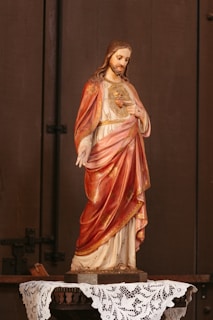 A statue of a religious figure with long hair and a beard, wearing a detailed robe adorned with golden patterns. The robe is a mix of rich red and white colors. The figure has one hand raised and the other touching its chest. The statue is placed on a table covered with a lace cloth, against a wooden background.