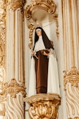 A statue of a nun is set in an ornate alcove with intricate gold detailing. The figure is dressed in a brown and white habit, holding a book in one hand and a religious artifact in the other. The sculpture is adorned with a metal crown or halo.