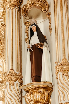 A statue of a nun is set in an ornate alcove with intricate gold detailing. The figure is dressed in a brown and white habit, holding a book in one hand and a religious artifact in the other. The sculpture is adorned with a metal crown or halo.