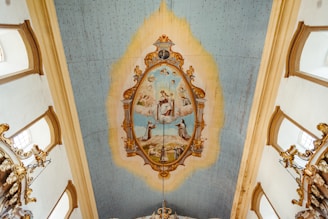 The image depicts an ornate ceiling with a central painting surrounded by an elaborate gold frame. The painting features several human figures set in a celestial scene with clouds and sky, possibly depicting religious or historical figures. The ceiling is adorned with light blue and cream tones, with decorative gold elements extending from the painting.