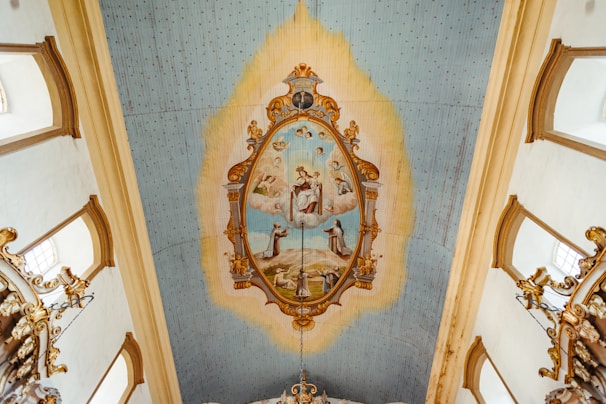 The image depicts an ornate ceiling with a central painting surrounded by an elaborate gold frame. The painting features several human figures set in a celestial scene with clouds and sky, possibly depicting religious or historical figures. The ceiling is adorned with light blue and cream tones, with decorative gold elements extending from the painting.