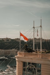 A red and white flag is prominently displayed on a building terrace overlooking a marina with numerous yachts. Behind the marina, a hilly landscape with several buildings is visible under a partly cloudy sky.
