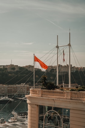 A red and white flag is prominently displayed on a building terrace overlooking a marina with numerous yachts. Behind the marina, a hilly landscape with several buildings is visible under a partly cloudy sky.