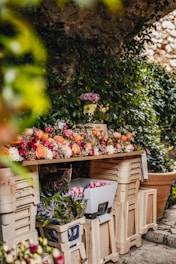 A wooden table covered with a variety of colorful flowers, including roses and tulips. Surrounding the table are several beige crates filled with additional flowers and foliage. The backdrop is lush with green ivy and stone walls, creating a serene, natural setting.