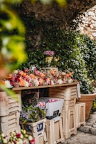 A wooden table covered with a variety of colorful flowers, including roses and tulips. Surrounding the table are several beige crates filled with additional flowers and foliage. The backdrop is lush with green ivy and stone walls, creating a serene, natural setting.