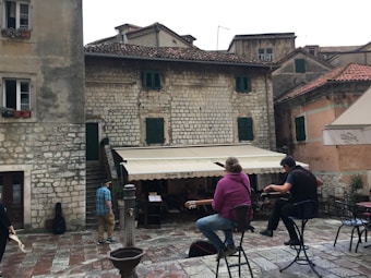 A rustic stone building with green shutters is the backdrop for an outdoor café scene. Two musicians, one playing a guitar, sit on stools performing to a small audience. A passerby in a plaid shirt and hat walks in the foreground. The cobblestone pavement adds to the historic atmosphere.