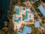 An aerial view of a resort featuring multiple swimming pools with surrounding lounge chairs and umbrellas arranged in a geometric pattern. The area is bordered by lush greenery and pathways leading to various buildings and structures.