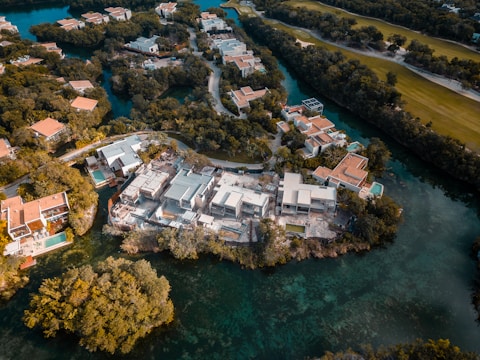 An aerial view of a residential area featuring modern houses with red-tiled roofs, surrounded by lush green trees and waterways. The landscape includes a prominent golf course, creating a scenic and upscale neighborhood environment.