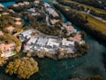 An aerial view of a residential area featuring modern houses with red-tiled roofs, surrounded by lush green trees and waterways. The landscape includes a prominent golf course, creating a scenic and upscale neighborhood environment.