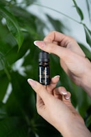 A close-up of hands holding a small, dark bottle labeled 'Crystal Weed' with a cannabis leaf logo. The background consists of large, lush green leaves.