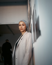 A woman stands in a gallery space, leaning against a wall lined with photographs. She wears a light headscarf and a beige coat over a dark top. The setting has a modern, minimalist design with soft lighting, creating a calm and introspective atmosphere.