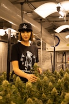 A person stands in an indoor cultivation facility surrounded by cannabis plants. They are wearing a black hat with a leaf design and a black t-shirt featuring a printed graphic. The environment is illuminated by hanging lights, and ventilation ductwork is visible above.