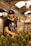 A person stands in an indoor cultivation facility surrounded by cannabis plants. They are wearing a black hat with a leaf design and a black t-shirt featuring a printed graphic. The environment is illuminated by hanging lights, and ventilation ductwork is visible above.