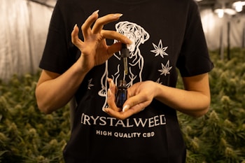 A person is holding a small bottle with a dropper in their hands. The person is wearing a black t-shirt featuring a design with a dog and cannabis leaves, along with the text 'CRYSTALWEED' and 'HIGH QUALITY CBD'. In the background, there are rows of cannabis plants, suggesting an indoor growing facility.