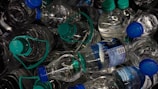 A pile of plastic water bottles with blue and green caps, some of which have labels with branding and information. The bottles are tightly packed together, creating a chaotic and cluttered appearance.