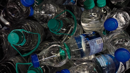 A pile of plastic water bottles with blue and green caps, some of which have labels with branding and information. The bottles are tightly packed together, creating a chaotic and cluttered appearance.