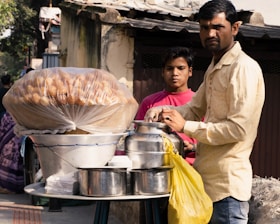 A street vendor is preparing or serving food beside a makeshift stall. The stall has a large container covered with plastic, holding food items that look like round fried snacks. There are jars, pots, and other utensils placed on the table, along with a yellow plastic bag. The vendor wears a light-colored shirt and is accompanied by a young boy in a pink shirt.