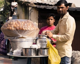 A street vendor is preparing or serving food beside a makeshift stall. The stall has a large container covered with plastic, holding food items that look like round fried snacks. There are jars, pots, and other utensils placed on the table, along with a yellow plastic bag. The vendor wears a light-colored shirt and is accompanied by a young boy in a pink shirt.