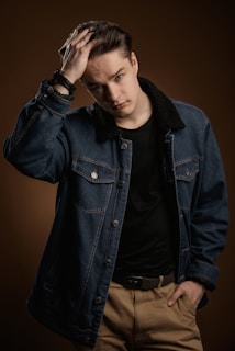A person is wearing a denim jacket with a black shirt underneath and khaki pants, holding their hair with one hand and posing against a brown backdrop. The look combines casual and stylish elements.