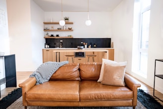 A modern living space featuring a tan leather couch with two pillows, one white and one beige, and a black-and-white striped throw draped over one side. Behind the couch is a minimalistic kitchen with light wood cabinetry, black tile backsplash, and open wooden shelves displaying various kitchen items. The room is well-lit by natural light coming through a large window, with two pendant lights hanging over the kitchen area.
