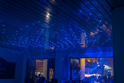 A room is illuminated with blue laser lights creating a dynamic and scattered pattern across the ceiling. The light interacts with the space, casting vibrant beams and reflections. The environment appears to be lively, with the light creating a futuristic or party-like atmosphere. Through an open doorway, a warmly lit interior of another space can be seen, suggesting a contrast between the areas.