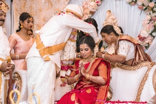 A bride in a red sari, adorned with jewelry, is seated and being blessed in a ceremonial setting. She is surrounded by people in traditional attire, including a person standing and placing a flower garland around her. The background includes floral decorations and ornate furniture.