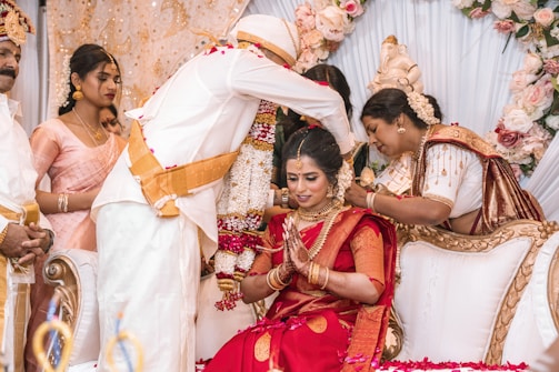 A bride in a red sari, adorned with jewelry, is seated and being blessed in a ceremonial setting. She is surrounded by people in traditional attire, including a person standing and placing a flower garland around her. The background includes floral decorations and ornate furniture.