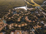 An aerial view of a lush, green landscape featuring a network of waterways interspersed with numerous buildings, most of which have brown roofs. The area is densely populated with trees, creating a natural and serene environment. The water reflects some sunlight, adding a dynamic quality to the scene.