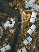 Aerial view of modern buildings surrounded by lush greenery and a river or waterway cutting through the landscape. The architecture is angular with flat roofs, and the buildings are interspersed with trees and pathways. The setting appears peaceful, with a blending of nature and urban design.