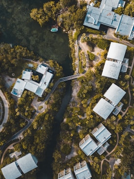 Aerial view of modern buildings surrounded by lush greenery and a river or waterway cutting through the landscape. The architecture is angular with flat roofs, and the buildings are interspersed with trees and pathways. The setting appears peaceful, with a blending of nature and urban design.
