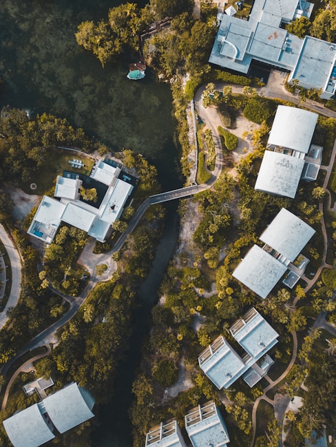 Aerial view of modern buildings surrounded by lush greenery and a river or waterway cutting through the landscape. The architecture is angular with flat roofs, and the buildings are interspersed with trees and pathways. The setting appears peaceful, with a blending of nature and urban design.