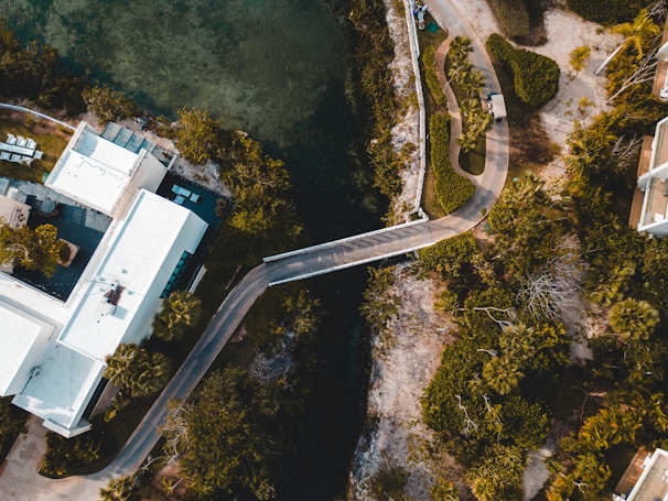 An aerial view of a modern building complex with white rooftops, surrounded by lush green trees. A narrow, winding road passes through the landscape, leading to a bridge that crosses over a waterway. The scene is a mix of natural and urban elements.