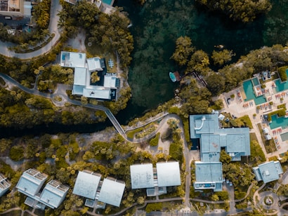 An aerial view featuring modern buildings surrounded by lush greenery. A river or water body runs through the landscape, and the buildings have angular, geometric shapes. There are rooftops with some visible infrastructure and pathways connecting different structures. A swimming pool area can be seen on the right with blue water and a few scattered lounging chairs.