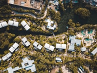 Aerial view of a resort complex with multiple white-roofed buildings surrounded by lush greenery. The layout is organized with winding pathways and a body of water at the top. There are recreational areas, likely with swimming pools, and a mix of natural and built environments.