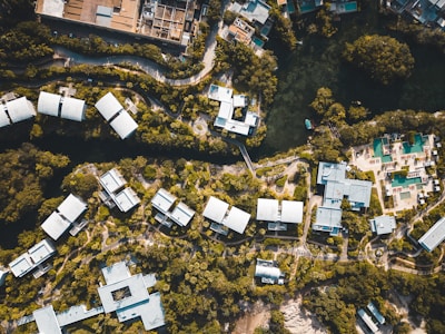 Aerial view of a resort complex with multiple white-roofed buildings surrounded by lush greenery. The layout is organized with winding pathways and a body of water at the top. There are recreational areas, likely with swimming pools, and a mix of natural and built environments.