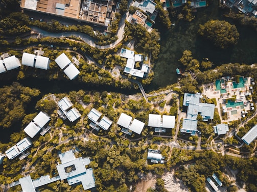 Aerial view of a resort complex with multiple white-roofed buildings surrounded by lush greenery. The layout is organized with winding pathways and a body of water at the top. There are recreational areas, likely with swimming pools, and a mix of natural and built environments.