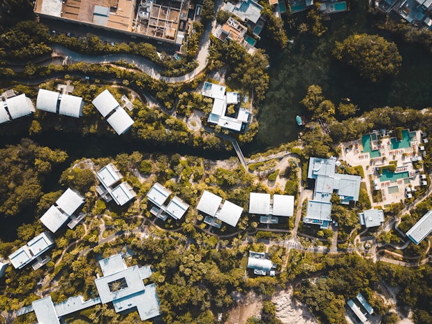 Aerial view of a resort complex with multiple white-roofed buildings surrounded by lush greenery. The layout is organized with winding pathways and a body of water at the top. There are recreational areas, likely with swimming pools, and a mix of natural and built environments.