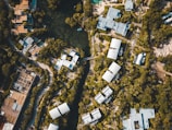 An aerial view of a residential area surrounded by dense greenery. Clustered modern buildings with light-colored roofs are interconnected by winding roads and pathways. A central water body is visible, encircled by trees, adding a natural element to the urban landscape.