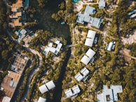 An aerial view of a residential area surrounded by dense greenery. Clustered modern buildings with light-colored roofs are interconnected by winding roads and pathways. A central water body is visible, encircled by trees, adding a natural element to the urban landscape.