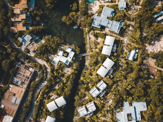 An aerial view of a residential area surrounded by dense greenery. Clustered modern buildings with light-colored roofs are interconnected by winding roads and pathways. A central water body is visible, encircled by trees, adding a natural element to the urban landscape.