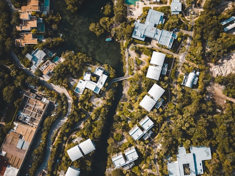 An aerial view of a residential area surrounded by dense greenery. Clustered modern buildings with light-colored roofs are interconnected by winding roads and pathways. A central water body is visible, encircled by trees, adding a natural element to the urban landscape.