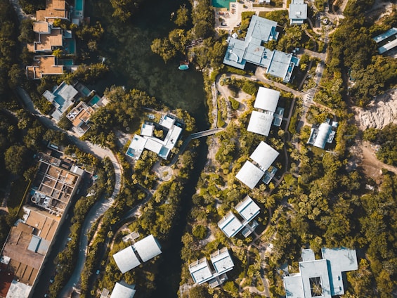 An aerial view of a residential area surrounded by dense greenery. Clustered modern buildings with light-colored roofs are interconnected by winding roads and pathways. A central water body is visible, encircled by trees, adding a natural element to the urban landscape.