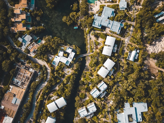 An aerial view of a residential area surrounded by dense greenery. Clustered modern buildings with light-colored roofs are interconnected by winding roads and pathways. A central water body is visible, encircled by trees, adding a natural element to the urban landscape.