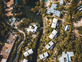 An aerial view of a residential area surrounded by dense greenery. Clustered modern buildings with light-colored roofs are interconnected by winding roads and pathways. A central water body is visible, encircled by trees, adding a natural element to the urban landscape.