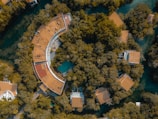 Aerial view of a circular building with a large roof partially covered by greenery, surrounded by dense green trees and other smaller buildings with similar roofs. The structures are set within a forested area interspersed with water bodies.
