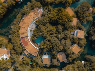 Aerial view of a circular building with a large roof partially covered by greenery, surrounded by dense green trees and other smaller buildings with similar roofs. The structures are set within a forested area interspersed with water bodies.