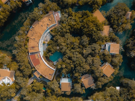 Aerial view of a circular building with a large roof partially covered by greenery, surrounded by dense green trees and other smaller buildings with similar roofs. The structures are set within a forested area interspersed with water bodies.