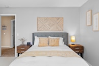 A modern bedroom features a gray upholstered headboard and a neatly made bed with white linens and mustard-colored accents. Two wooden nightstands flank the bed, each with a small lamp and decorative items. A patterned artwork hangs above the headboard, and two framed artworks are displayed on the adjacent wall. The room has a neutral color palette with gray walls and beige carpet.