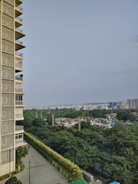 A tall, multi-storey building with balconies on the left side overlooks a cityscape filled with lush green trees. In the distance, numerous other buildings, both low and high rise, are visible under a clear blue sky. The area appears residential with greenery and urban elements blending harmoniously.